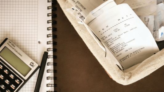 imagem de um caderno com uma calculadora e lápis em cima e do outro lado as notas fiscais. tudo em cima de uma mesa.